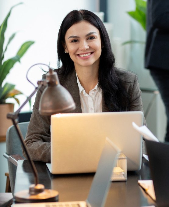 Smiling woman sitting at a desk with a laptop, wearing a business suit. She is in a modern office environment with plants and a lamp on her desk.