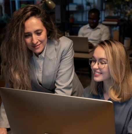 Two women working together at a desk, looking at a computer screen. One woman is standing and the other is seated, both smiling and focused. The office setting has a warm, professional atmosphere with another colleague working in the background.