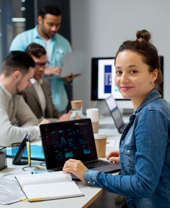 Young woman sitting at a desk with a laptop displaying data and graphs, smiling at the camera. She is in a modern office environment with several colleagues working in the background, collaborating on laptops and discussing work.