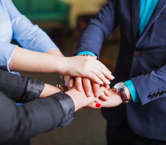 Close-up of multiple hands stacked together in a gesture of teamwork and unity. The individuals are dressed in professional attire, symbolizing collaboration and partnership in a business setting.