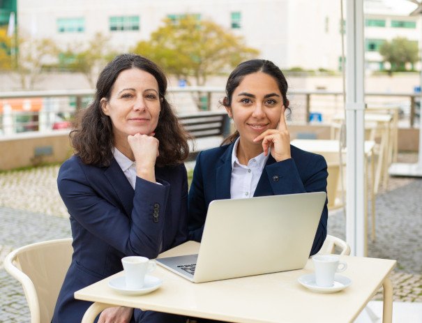 Two businesswomen sitting at an outdoor café table with a laptop and coffee cups. Both are dressed in professional attire, looking confidently at the camera, with a relaxed and collaborative atmosphere in an urban setting.