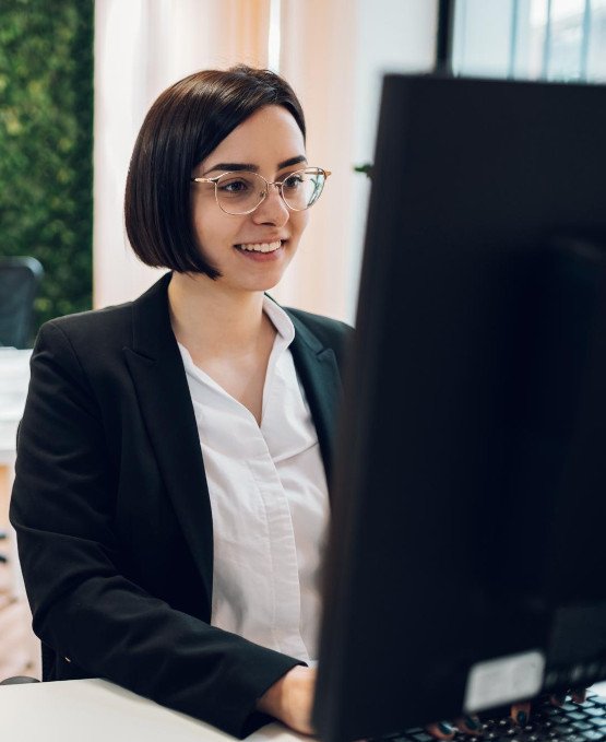 Young woman in a professional setting, wearing glasses and a business suit, working on a desktop computer. She is smiling and appears focused, sitting in a modern office with natural lighting and a green plant wall in the background.