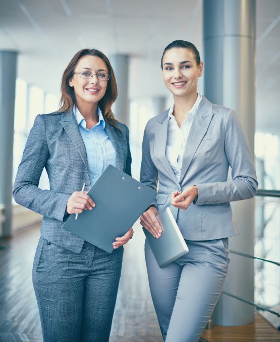 Two professional women standing in a modern office hallway, both dressed in business suits. One is holding a clipboard, and the other has a tablet, smiling confidently at the camera. The setting features large windows and a bright, spacious atmosphere.