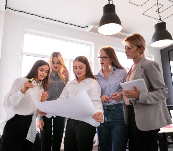 Group of five women in a modern office collaborating on a project. They are gathered around a large sheet of paper, discussing and analyzing the details. The atmosphere is focused, with one woman holding a tablet and others using markers to highlight key points.