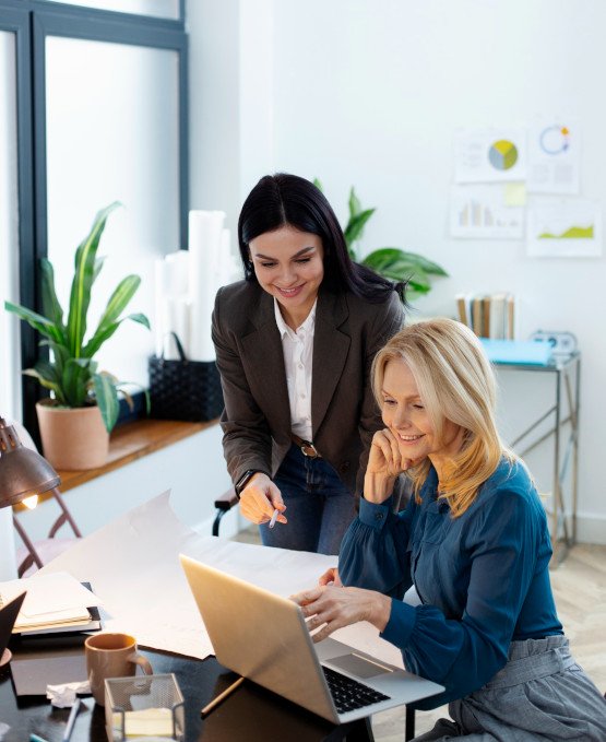 Two women in a bright office working together at a desk. One woman is seated, looking at a laptop and smiling, while the other is standing beside her, pointing at the screen. The setting includes plants, charts on the wall, and work-related documents on the desk, creating a collaborative and productive environment.