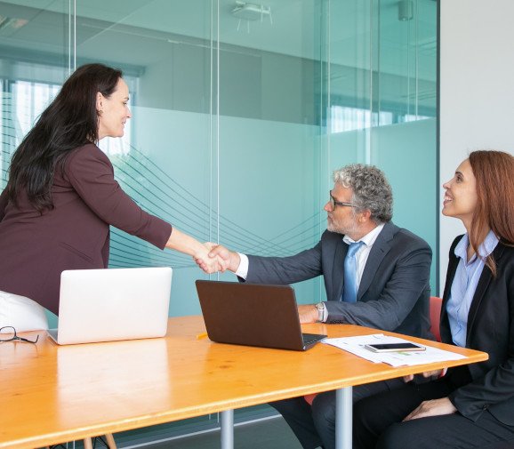 Professional business meeting in a modern office. A woman is standing and shaking hands with a seated man across a table, while another woman sits beside him, smiling. Laptops and documents are on the table, indicating a formal discussion or agreement.