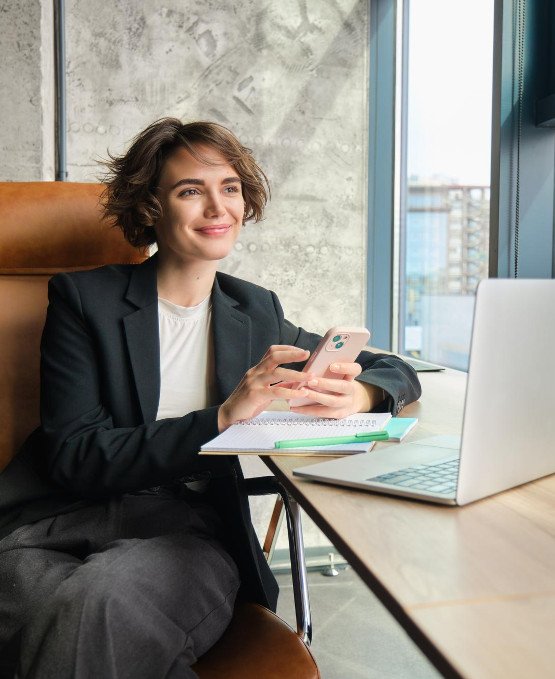 Young woman sitting in a modern office, holding a smartphone and smiling while looking out the window. She has a notebook and laptop on the desk in front of her, creating a relaxed yet professional work environment.