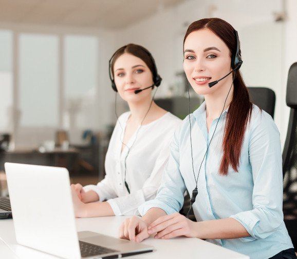 Two female customer service representatives sitting at a desk, wearing headsets and smiling. They are working in a bright, modern office environment with laptops in front of them, ready to assist clients.